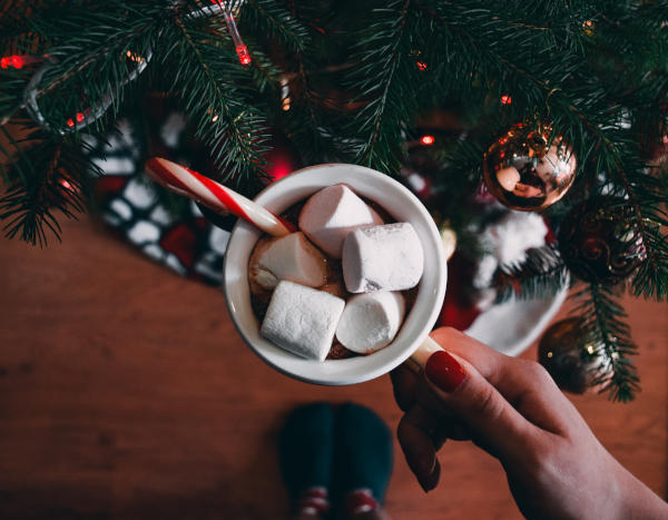 Jemand hält eine Tasse mit Kakao und einer Zuckerstange und Marshmallows darin vor einem Weihnachtsbaum, Blick direkt in die Tasse.